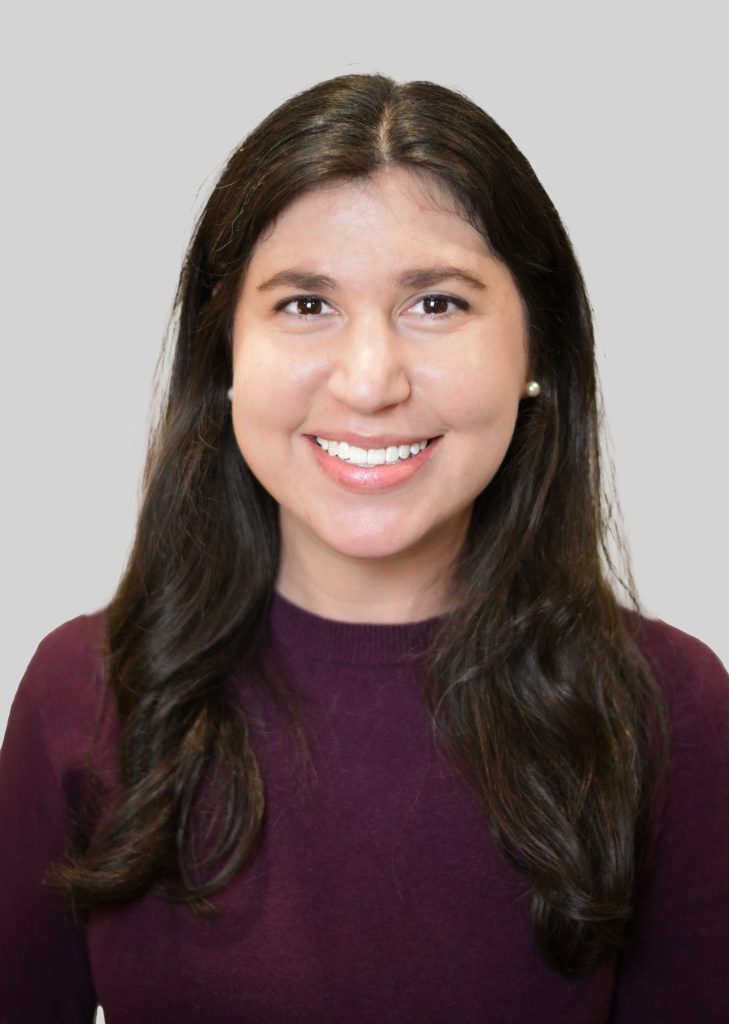 Young therapist Jona Koplow in a dark purple sweater, smiling against a gray background at Serengeti Wellness.