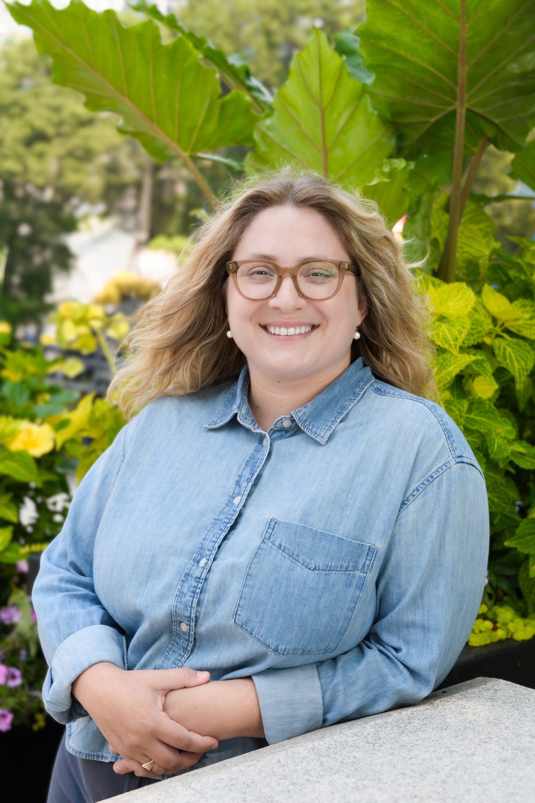 Laura Johnson, Administrative Assistant at Serengeti Wellness, smiling outdoors with lush green plants and flowers in the background, wearing a denim shirt.