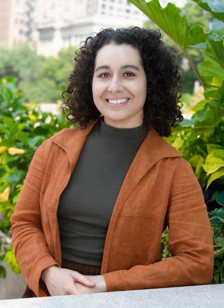 Natalia Gurmendi, therapist at Serengeti Wellness, smiling in a green outdoor setting with vibrant foliage, wearing an orange jacket and a dark top, embodying a welcoming and supportive atmosphere for anxiety therapy.
