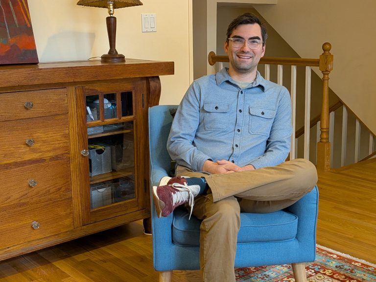 Man sitting comfortably in a blue armchair, smiling, in a cozy therapy office setting with wooden furniture and warm lighting, reflecting a supportive environment for discussing men's mental health.