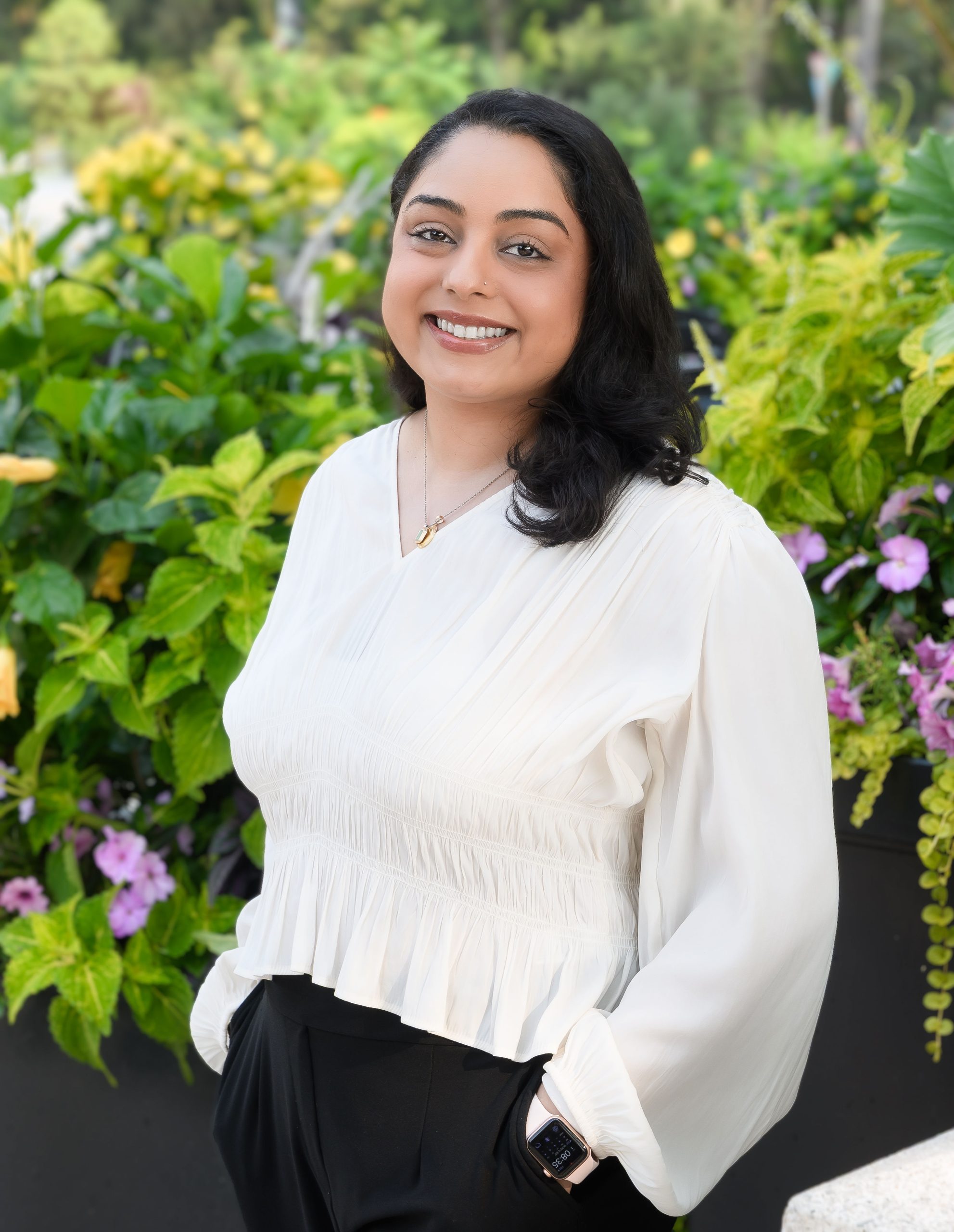 Sanya Jaffer, Clinic Operations Manager at Serengeti Wellness, smiling in a white blouse surrounded by lush greenery and flowers, emphasizing community and support in mental health services.