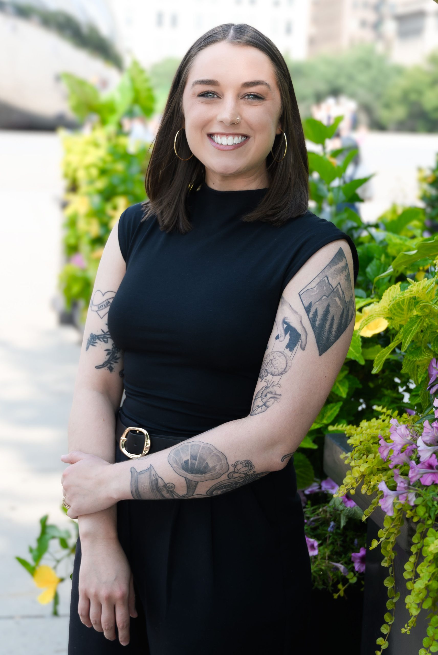 Smiling woman in black sleeveless top with tattoos, standing in front of vibrant greenery, representing the diverse team at Serengeti Wellness, a mental health therapy provider in Naperville.