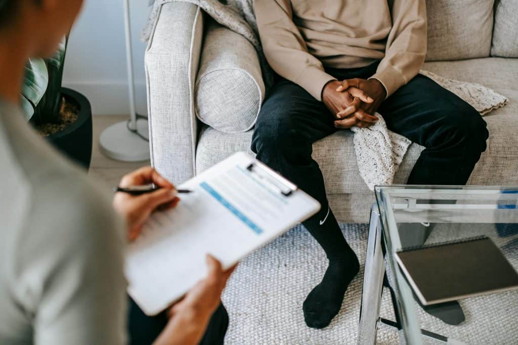 Client seated on a couch during a therapy session, with a therapist taking notes on a clipboard, emphasizing emotional support and recovery in a compassionate setting.