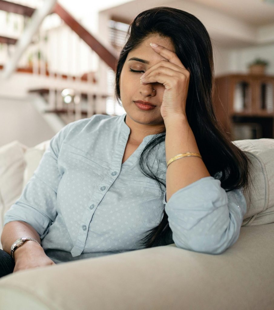Woman sitting on a couch, holding her forehead in a gesture of stress or fatigue, symbolizing anxiety and mental health challenges in a home setting.