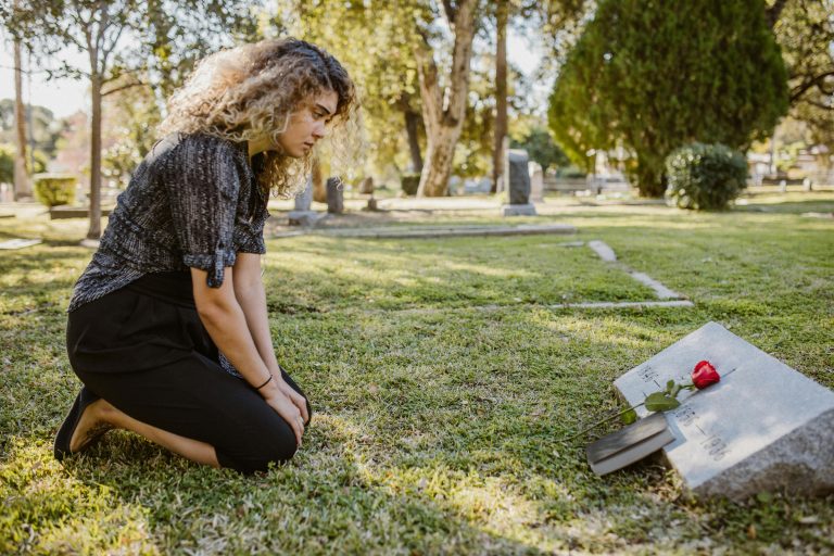 Woman kneeling at a grave with a red rose and a book, reflecting on loss in a cemetery, symbolizing grief and remembrance.