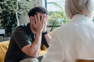 Man with hands covering face in therapy session, expressing emotional distress, seated on couch, with therapist in white attire, indoor setting with greenery, reflecting mental health support at Serengeti Wellness.